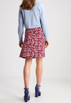Femme portant un pull court bleu clair, une jupe rouge à fleurs et des chaussures à talons bleues, debout de dos sur un fond uni.