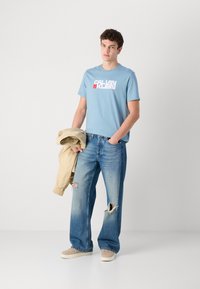 Šviesiai mėlynas medvilninės medžiagos marškinėliai su "CALVIN KLEIN" logotipu, derinami su šviesiai mėlynomis nudilusiomis džinsais ir smėlio spalvos pūkuotu švarku, laikomu rankoje.