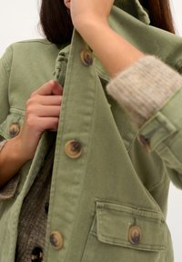 Person wearing a beige sweater buttoning up a green jacket with large brown buttons against a plain background.
