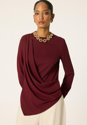 Femme portant un haut drapé bordeaux à manches longues, assorti à un pantalon crème, un collier chaîne doré épais et des boucles d’oreilles assorties.