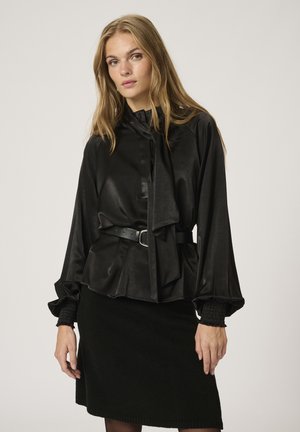 Camicia nera di satin con collo a cravatta, design plissettato e maniche lunghe a sbuffo. Abbinata a una gonna nera strutturata e una cintura.