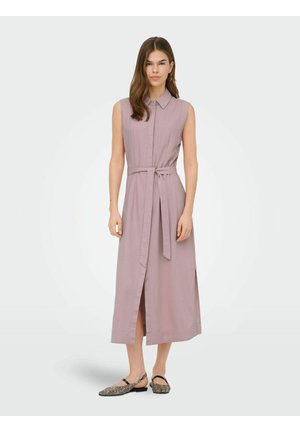 Nő áll, aki ujjatlan, levendulaszínű midi ruhát visel, gallérral, elöl hasítékkal és anyagövvel, mintás pántos lapos cipővel párosítva.