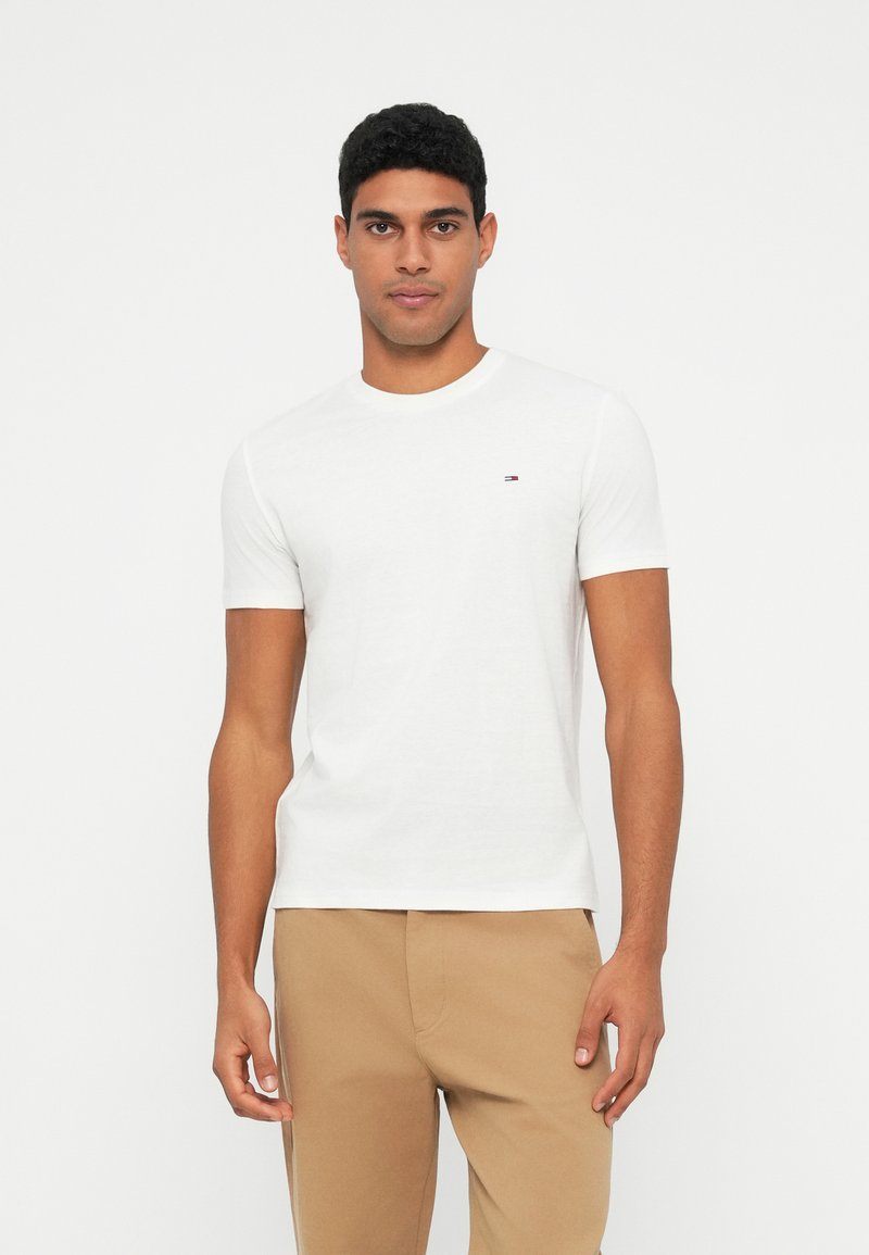 White cotton t-shirt with short sleeves and a round neckline; small logo detail on the left chest; paired with tan trousers.