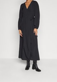 Svart omslagsklänning med långa puffärmar, v-ringning, midjeband och en svängd kjol, kombinerad med svarta klackstövlar. Slät tygtextur.