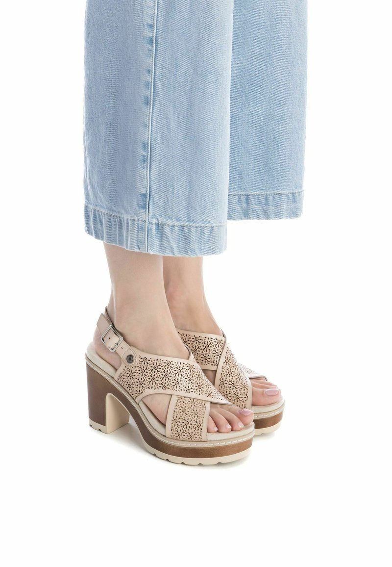 Sandalias de tacón alto perforadas de color beige con amplias correas entrelazadas, hebilla ajustable en el tobillo y suela texturizada para mayor agarre.