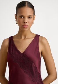 Bordó szatén ruha V-kivágással, elöl átlós fekete csipkehálóval, sima textúrával és ujjatlan kialakítással.