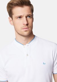 Biała koszulka polo o strukturalnym wykończeniu. Posiada kołnierzyk wykończony na niebiesko oraz zapięcie na guziki, z małym niebieskim logo na piersi.