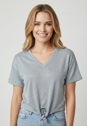 Donna sorridente con capelli biondi indossa una maglia argento scintillante a maniche corte con scollo a V annodata in vita e jeans azzurri su uno sfondo neutro.