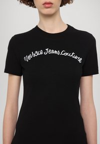 Melna T-krekls īsām piedurknēm ar „Versace Jeans Couture” izšuvumu baltā krāsā krūšu daļā. Klasiska apaļa apkakle.