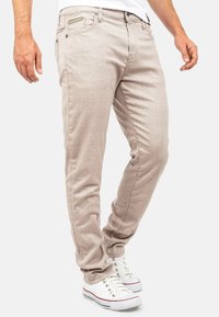 Jean beige slim fit avec des rayures verticales, design à cinq poches et détail de poche zippée. Présente une texture lisse et des coutures classiques.