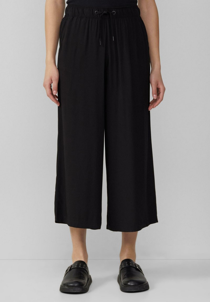 Zwarte wijde broek van zacht materiaal, met een elastische tailleband en een trekkoord, en zijsplitten voor gemakkelijkere beweging.
