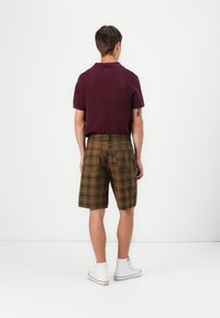Mandens rygview iført en bordeaux tekstureret polo skjorte og grøn-brune ternede shorts, parret med hvide højhælede sneakers.