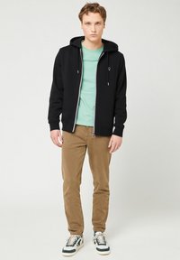 Schwarze Zip-Hoodie mit Kordelzugkapuze, darunter ein hellgrünes T-Shirt, kombiniert mit braunen Cordhosen und gemusterten Sneakers.