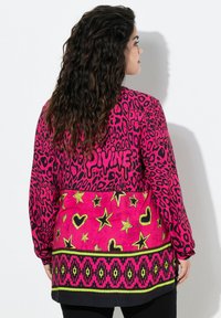 Vrouw met lang krullend haar die een felroze jack draagt met een zwart luipaardpatroon, gele sterren, harten en het woord "DIVINE" op de rug.