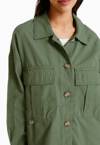 Olivgrön arbetsoverall med knappstängning fram, två bröstfickor och krage, tillverkad av lättviktig tyg med en mjuk textur.