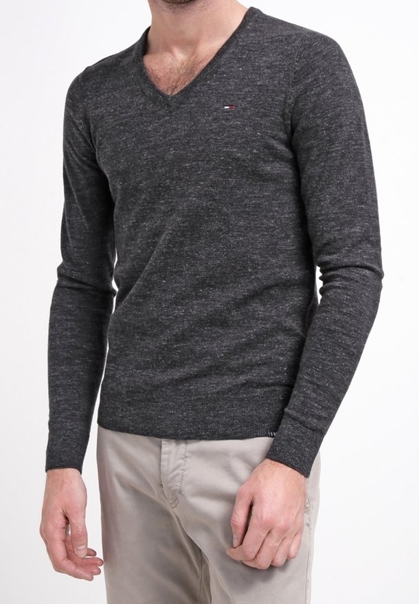 Homme portant un pull à col en V gris foncé avec un petit logo sur la poitrine et un pantalon beige, debout devant un fond blanc.