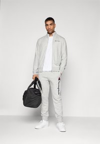 Grå hettejakke med glidelås og matchende joggebukser med brodert logo. Hvit t-skjorte under. Svart treningsbag. Hvite joggesko.