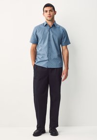 Chemise à manches courtes bleu clair avec un motif géométrique, boutonnée sur le devant, associée à un pantalon large noir et des chaussures noires.