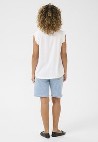 Top sans manches blanc avec des bords froufroutants, associé à un short en denim bleu moyen. Des chaussures plates noires complètent la tenue décontractée.