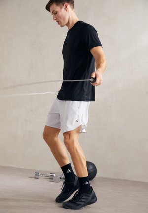 Schwarzes Sport-T-Shirt und weiße Shorts mit seitlichen Schlitzen. Die Person springt Seil und trägt schwarze Turnschuhe und Socken auf einem glatten Boden.