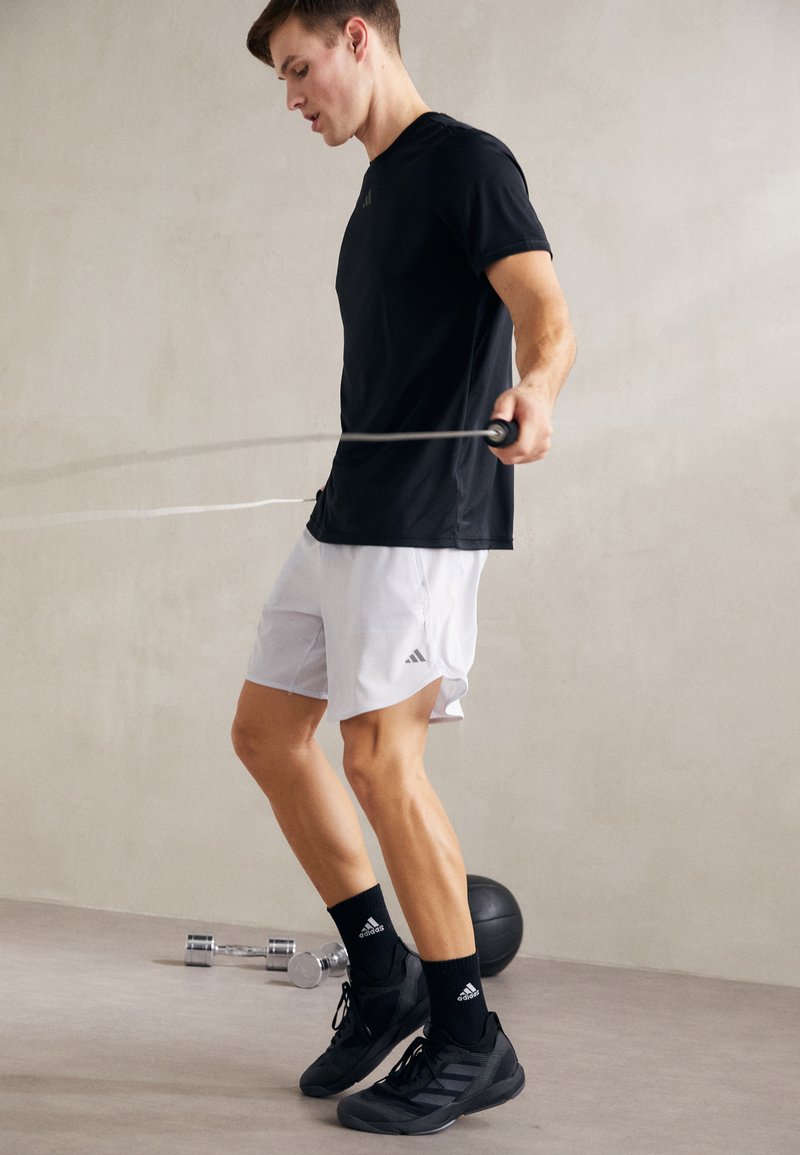 Camiseta atlética negra y pantalones cortos blancos con aberturas en los laterales. La persona está saltando la cuerda, vistiendo zapatillas y calcetines negros, sobre un suelo liso.