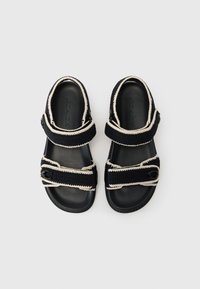 Black sandals with textured fabric straps, cream stitching, and adjustable buckles. Features a smooth black sole and a minimalistic design.