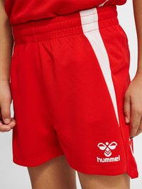 Enfant portant un short de sport rouge avec des bandes blanches sur les côtés et le logo hummel sur la partie inférieure de la jambe gauche, mains détendues sur les côtés.
