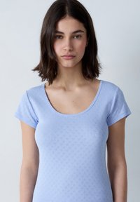 Camiseta de manga corta azul claro con escote redondeado, patrón texturizado y borde en forma de ola alrededor del escote. Diseño simple y ajustado.