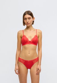 Červená krajková souprava spodního prádla obsahující braletku s vroubkovanými okraji a nastavitelnými ramínky, doplněná o kalhotky s krajkovým lemem.