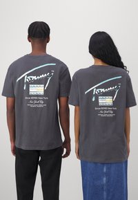 Camisetas de algodón gris con una gran impresión gráfica en la parte posterior. Presenta el logo de Tommy Jeans, texto y franjas horizontales multicolores.