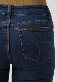 Temno modri denim hlače s prilegajočim dizajnom, z zlatim šivom in majhno črno oznako na zadnjem žepu.
