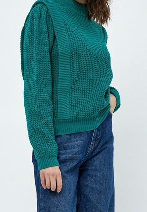 Mujer con suéter de punto texturizado en color teal con paneles acanalados verticales, combinado con jeans de mezclilla azul, una mano en el bolsillo y la otra relajada a un lado.