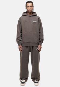 Grauer Kapuzenpullover mit Fronttasche und besticktem Text. Kombiniert mit passenden, locker sitzenden Jogginghosen. Weicher Stoff.
