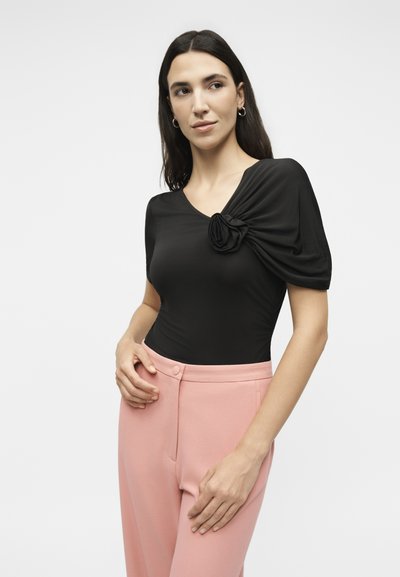 Γυναίκα με μακριά σκούρα μαλλιά που φοράει μαύρο τοπ με σατινέ φουσκωτό μανίκι και λουλουδένια λεπτομέρεια, συνδυασμένο με ψηλόμεσο ροζ παντελόνι, στεκόμενη μπροστά σε λευκό φόντο.