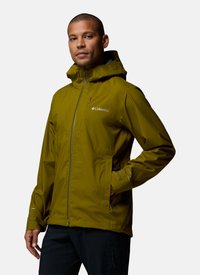 Man wearing olive green Columbia rain jacket with hood, zipped front, and pockets, standing with one hand in pocket against white background.
