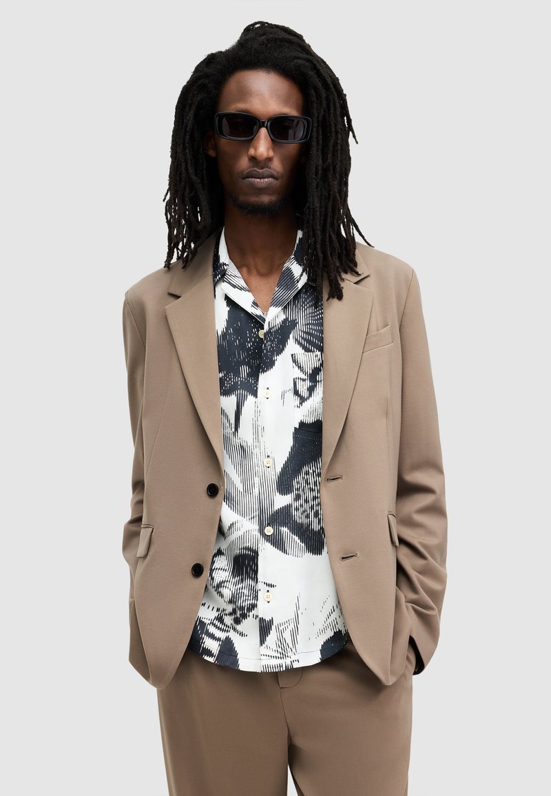 Chaqueta de traje marrón claro a medida con una camisa blanca y negra de patrón debajo. Los accesorios incluyen unas gafas de sol negras estrechas.