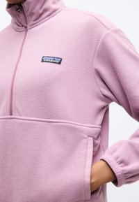 Φωτεινή μωβ ζακέτα Patagonia από φλις με φερμουάρ μπροστά και χέρι βυθισμένο στην πλαϊνή τσέπη.