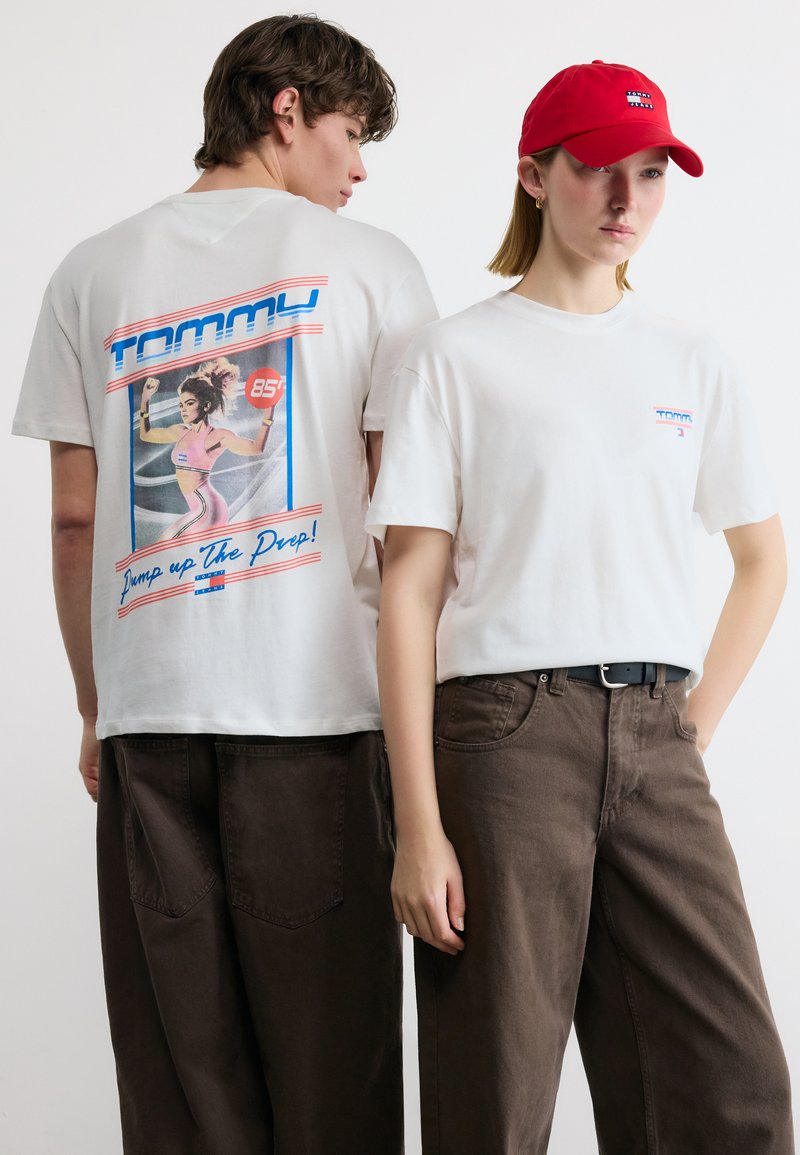 Δυο άτομα με λευκά t-shirts Tommy και σκούρα παντελόνια· το ένα φοράει κόκκινο καπέλο, ενώ η πλάτη του άλλου δείχνει ένα γραφικό με μια γυναίκα να ασκείται.