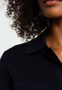 Marineblauwe gebreide kleding met een kraaghalslijn, voorzien van een ribbeltextuur en subtiele stikdetails. Close-up van het bovenste deel van de stof.