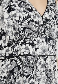 Černobílá tkanina s květinovým vzorem, zahrnující různé květiny a listy, s výstřihem do V a švem v pase, ozdobena černými lemovkami.