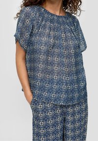 Blouse bleue transparente avec un motif géométrique agrémenté d'accents floraux. Encolure froncée et manches courtes, associée à un pantalon assorti au motif similaire.