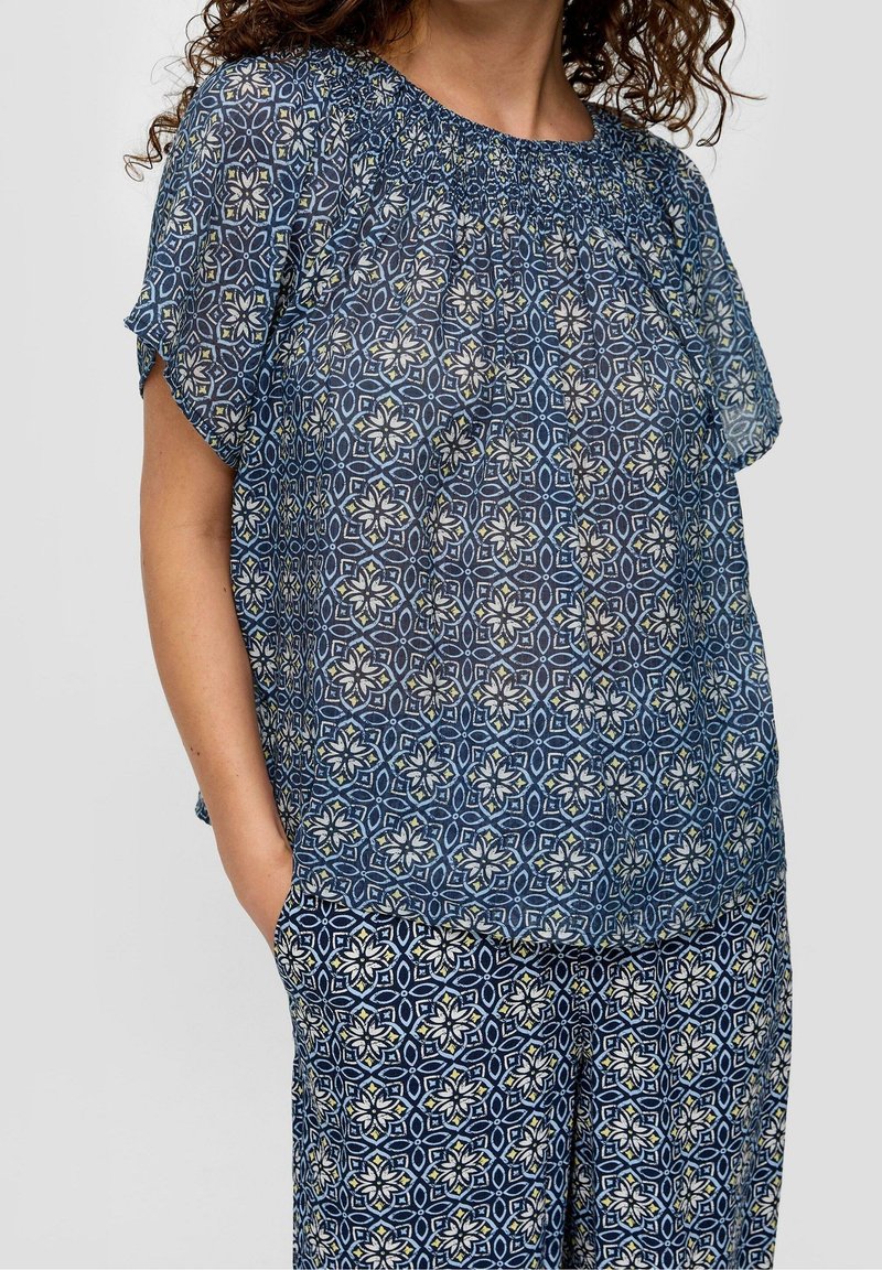 Blouse bleue transparente avec un motif géométrique agrémenté d'accents floraux. Encolure froncée et manches courtes, associée à un pantalon assorti au motif similaire.