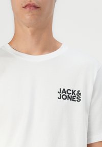 Hvit bomulls t-skjorte med rund hals og korte ermer. Har en svart "JACK & JONES" logo trykt på venstre bryst.