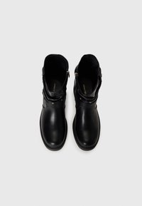 Paire de bottines noires avec fermeture éclair latérale et détail de sangle, vues de dessus sur fond gris clair.