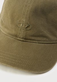 Gorra verde oliva hecha de tela acanalada, con un detalle de logo bordado y visera curva, con costuras visibles alrededor de los bordes.
