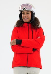 Kvinde iført en beige skihjelm med goggles, rød skijakke og lysegrå bukser, stående med armene korslagte mod en hvid baggrund.