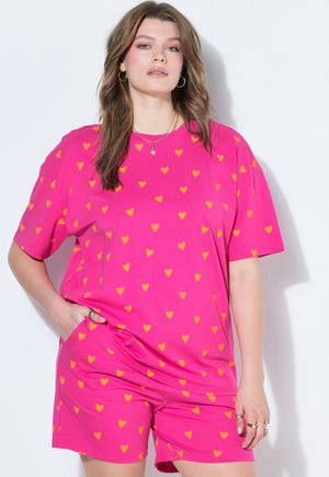 Femme portant un t-shirt et un short assortis rose vif avec un petit motif de cœurs orange, debout avec une main dans la poche.