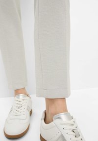 Pantalons gris clair en tissu texturé, avec une coupe droite et des bords ourlés, associés à des baskets blanches avec des accents métalliques.