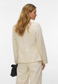 Femme aux cheveux blonds longs et ondulés portant une veste crème et un pantalon assorti, tenant une petite pochette noire dans sa main droite.