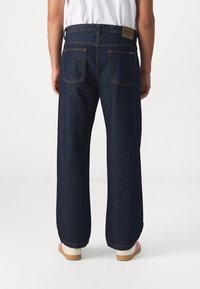 Jean en denim bleu foncé avec une coupe décontractée, présentant des coutures visibles, des accessoires en métal argenté, et une étiquette en cuir rectangulaire sur la taille.